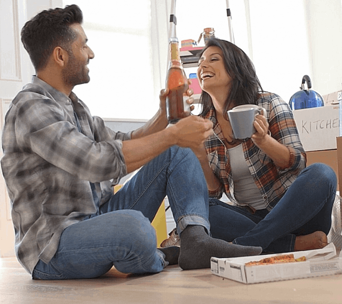 A Hispanic couple in their new home enjoying champagne and pizza on the floor surrounded by moving boxes