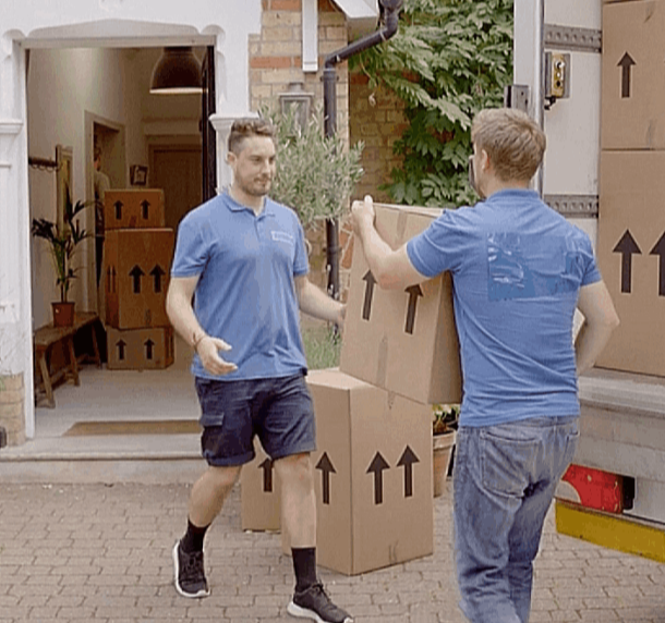 Movers in blue shirts unload moving boxes from a truck into a house